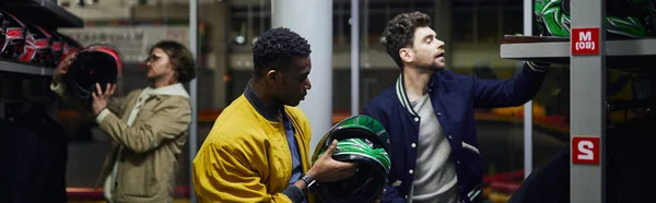 Three interracial men in casual clothes choosing helmets for karting, go-cart concept, banner — Stock Photo