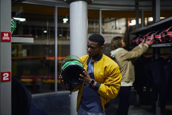 Uomo africano americano che sceglie il casco per il karting vicino amico su sfondo sfocato, concetto go-cart — Foto stock
