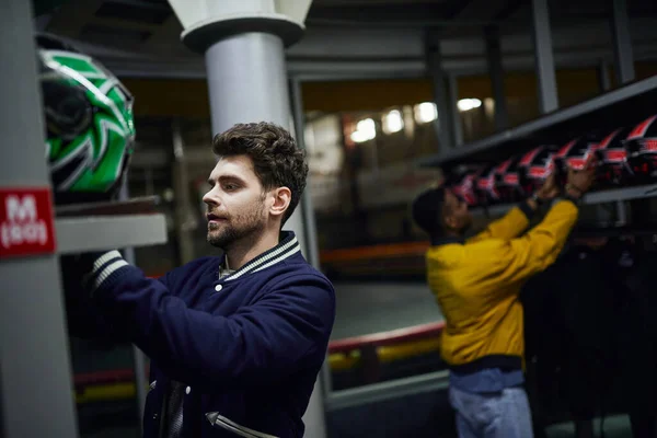 Handsome man choosing helmet for karting near african american friend on blurred backdrop, go-cart — Stock Photo