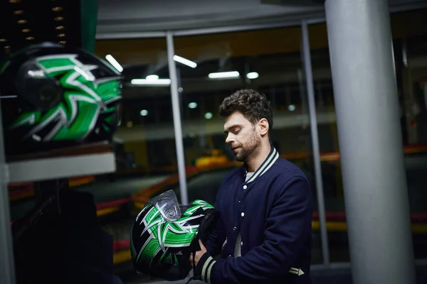 Hombre guapo que elige el casco para karting dentro del vestuario de karting, estantes con el engranaje del motor - foto de stock