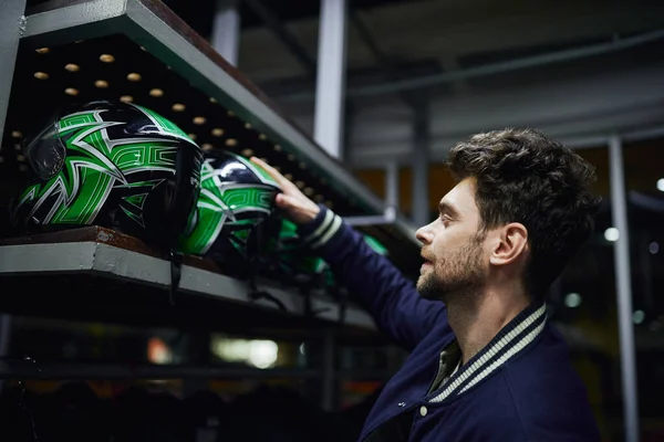 Side view of handsome man choosing helmet for karting inside of karting locker room, motorsport — Stock Photo