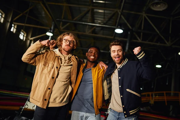 Three interracial male friends smiling and standing near racing cars inside of indoor karting track — Stock Photo