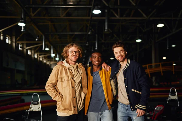 Amigos do sexo masculino multicultural sorrindo e de pé perto de carros de corrida dentro da pista de karting indoor — Fotografia de Stock