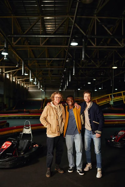 Amici maschi multiculturali sorridenti e in piedi vicino alle auto da corsa all'interno della pista di karting, sorridono — Foto stock