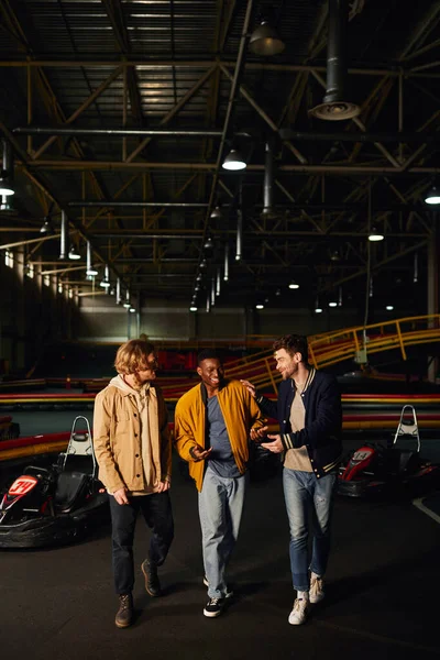 Amigos multiculturales sonriendo y charlando cerca de coches de carreras dentro de pista de karting, hobby y alegría - foto de stock