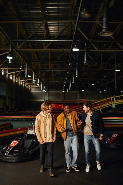 Cheerful diverse friends chatting and walking near racing cars inside of karting track, hobby — Stock Photo