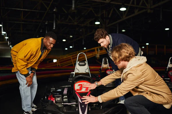 Amigos masculinos interracial felices mirando el karting rojo dentro de la pista cubierta, pasatiempo del automovilismo - foto de stock