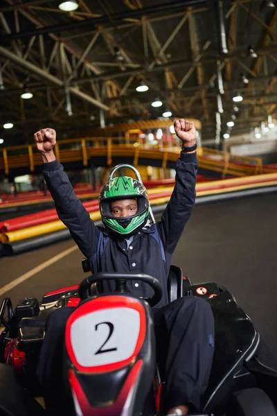 Go kart speed drive, thrilled african american driver in helmet raising hands and winning kart race — Stock Photo