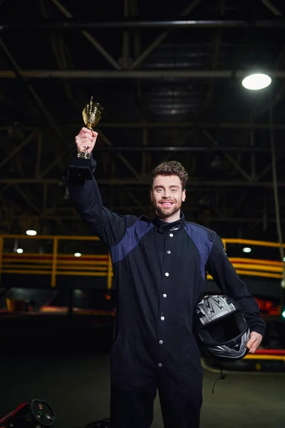 Allegro pilota in abbigliamento sportivo in possesso di coppa d'oro e in piedi come vincitore della gara go-kart, trofeo — Foto stock