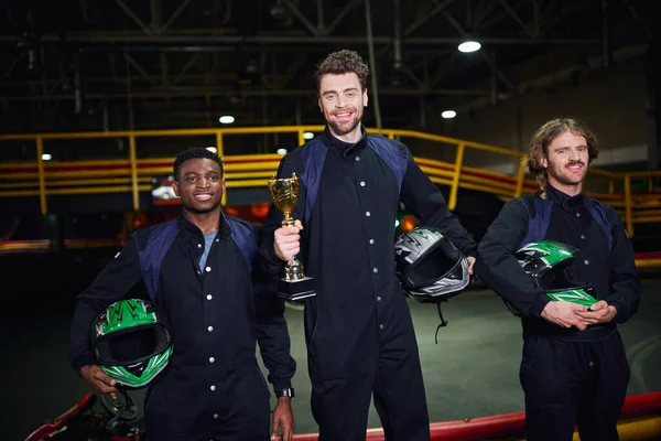 Happy driver holding golden cup and standing as a winner of go-kart race near diverse competitors — Stock Photo