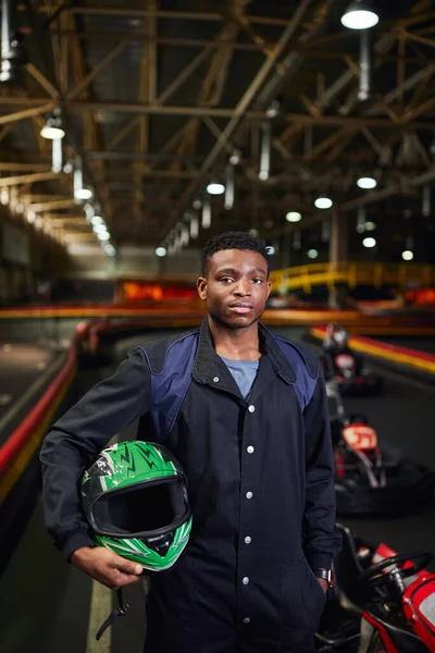 Hombre afroamericano en ropa deportiva sosteniendo casco y de pie en pista de karting cerca borrosa go-kart - foto de stock