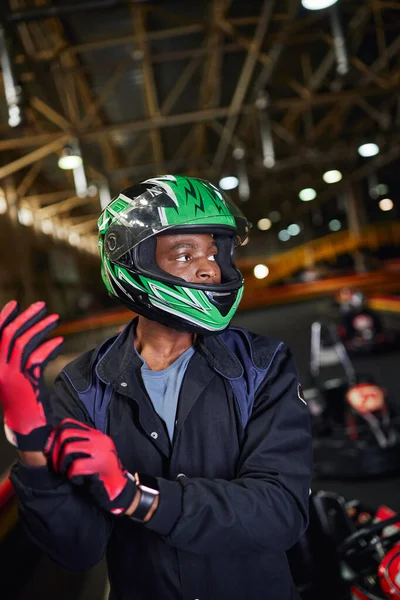 Pilota americano africano di sport motoristici in casco indossando guanti e in piedi in pista di karting — Foto stock