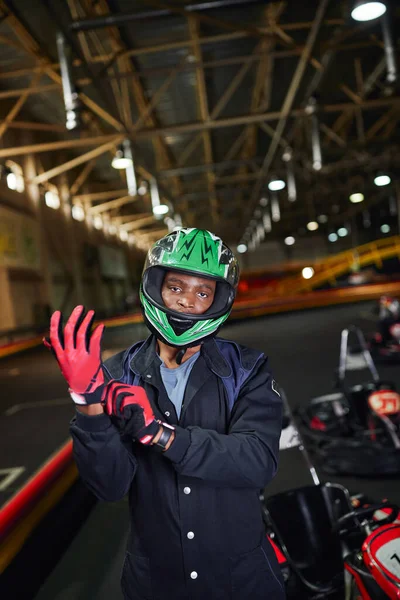 Ambitious african american motorsports driver in helmet wearing gloves and standing near go-kart — Stock Photo