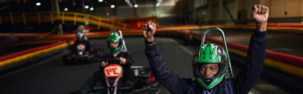 Aufgeregter afrikanisch-amerikanischer Go-Kart-Fahrer mit Helm, der Hände hebt und Rennen gewinnt, Go-Kart-Banner — Stockfoto