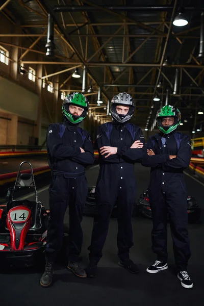 Trois coureurs de karting confiants en vêtements de sport et casques debout avec les bras croisés sur le circuit — Photo de stock