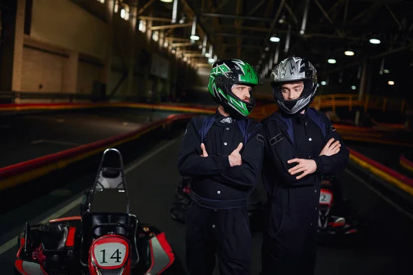 Deux coureurs de karting confiants en vêtements de sport et casques debout avec les bras croisés sur le circuit — Photo de stock