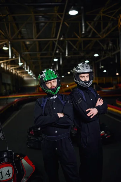 Two confident go kart drivers in sportswear and helmets standing with folded arms on circuit — Stock Photo