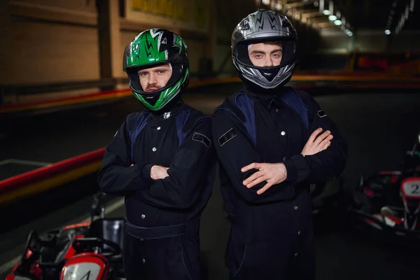Two confident go kart competitors in sportswear and helmets standing with folded arms on circuit — Stock Photo
