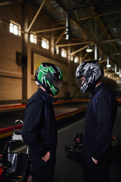 Dos competidores de karts en ropa deportiva y cascos de pie cara a cara en el circuito, karting interior - foto de stock