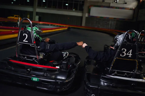Dos conductores de karts de ir en ropa deportiva y cascos puño chocando y sentado en coches deportivos para karting - foto de stock