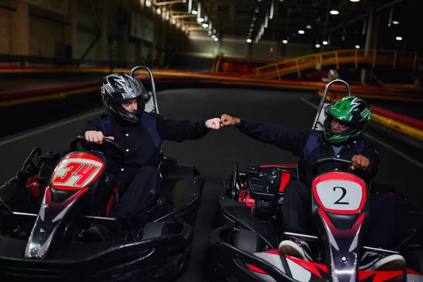 Diverse go kart drivers in helmets fist bumping and sitting in sport cars for karting on circuit — Stock Photo