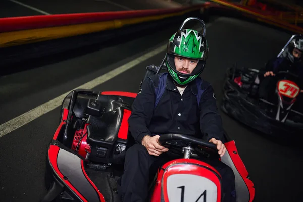 Uomo concentrato in abbigliamento sportivo e casco guida auto sportive per il karting vicino amico sul circuito coperto — Foto stock