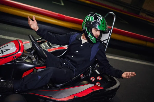 Conducteur émotionnel dans un geste de casque tout en conduisant une voiture de sport pour le karting sur le circuit intérieur — Photo de stock