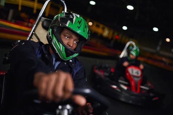 Africano americano uomo in casco guida go kart su circuito coperto vicino amico su sfondo sfocato — Foto stock