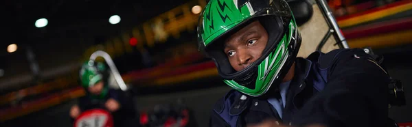 Africano americano hombre en casco conducción ir kart en circuito cerca amigo en borrosa telón de fondo, bandera - foto de stock