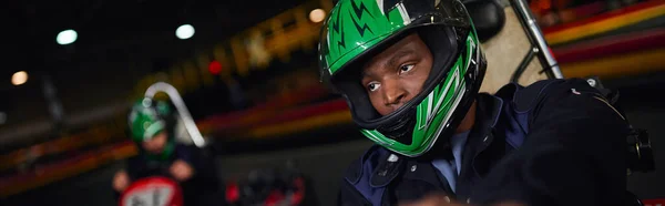 African american man in helmet driving go kart on circuit near friend on blurred backdrop, banner — Stock Photo