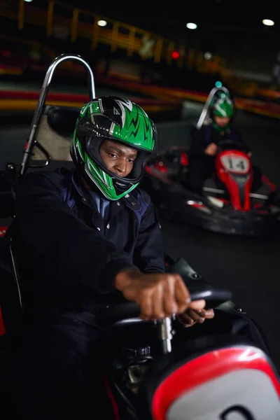 African american driver in helmet driving go kart on indoor circuit near friend on blurred backdrop — Stock Photo