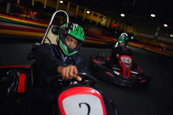 African american driver in helmet driving go kart on circuit near friend on blurred backdrop — Stock Photo