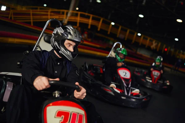 Concentrated man driving go kart near diverse drivers in helmets on indoor circuit, adrenaline — Stock Photo