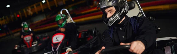 Concentrated man driving go kart near diverse drivers in helmets on indoor circuit, banner — Stock Photo