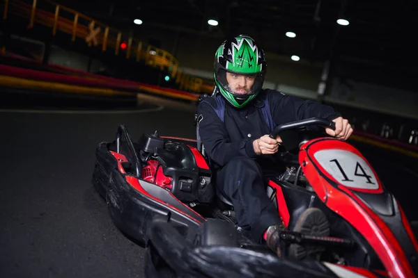 Emotionaler Rennfahrer in Helm und Sportbekleidung fährt Go-Kart auf Indoor-Rundkurs, Adrenalin-Konzept — Stockfoto