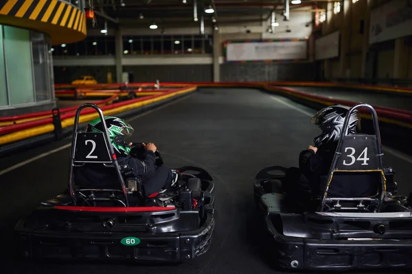 Dos diversos corredores en cascos y ropa deportiva conducción ir kart en circuito cubierto, competidores - foto de stock
