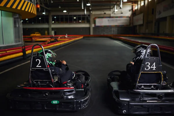 Dos diversos corredores en cascos y ropa deportiva conducción ir kart en circuito cubierto, competidores - foto de stock