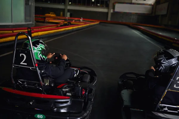 Dos hombres multiculturales en cascos y ropa deportiva conducción ir kart en circuito cubierto, competidores - foto de stock