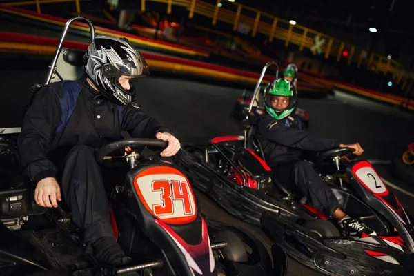 Competidores multiétnicos dirigindo ir kart no circuito interno, corrida de velocidade e automobilismo — Fotografia de Stock