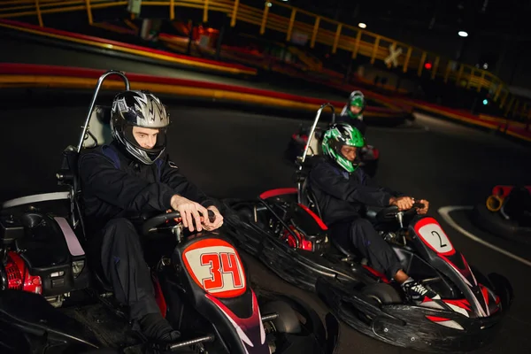 Les coureurs multiculturels concentrés conduisent un chariot de karting sur circuit intérieur, course de vitesse et sport automobile — Photo de stock