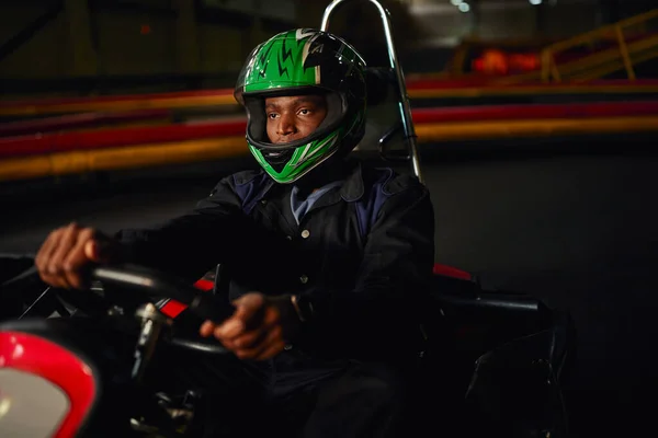 Pilota di go kart afroamericano in casco guida su circuito indoor, sfida di velocità — Foto stock