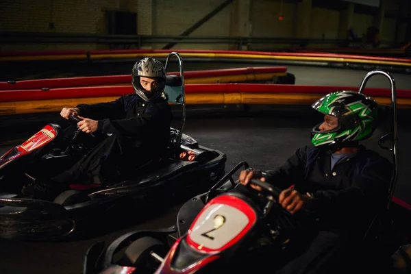 Aller pilote de kart dans le casque en regardant un concurrent afro-américain et la conduite sur circuit, la vitesse — Photo de stock