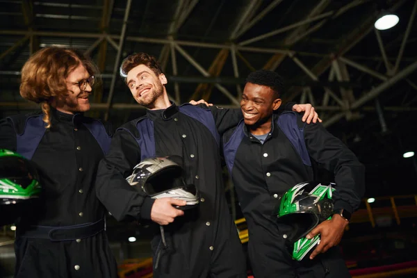Team of interracial and happy go kart drivers in protective suits hugging and holding helmets — Stock Photo