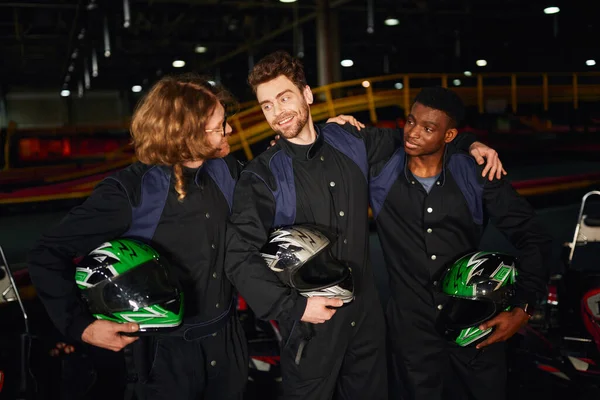 Group of interracial and happy go kart drivers in protective suits hugging and holding helmets — Stock Photo