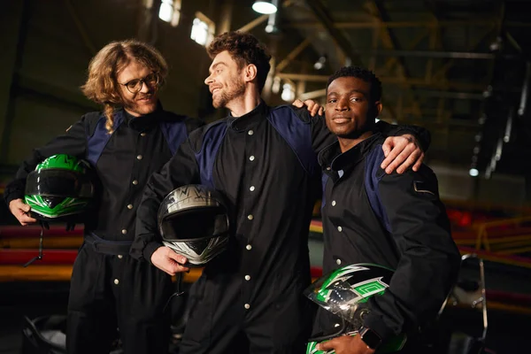 Group of multicultural and happy go kart drivers in protective suits hugging and holding helmets — Stock Photo