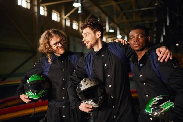 Group of multicultural and happy go kart racers in protective suits hugging and holding helmets — Stock Photo