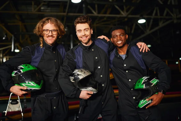 Group of multicultural and joyful go kart racers in protective suits hugging and holding helmets — Stock Photo