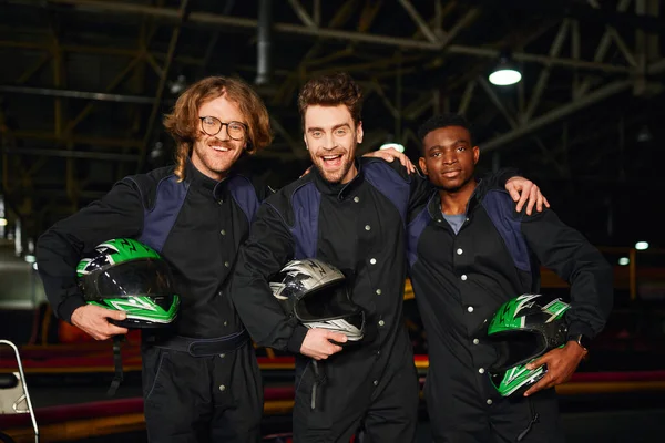 Multicultural and excited go kart racers in protective suits hugging and holding helmets, winners — Stock Photo