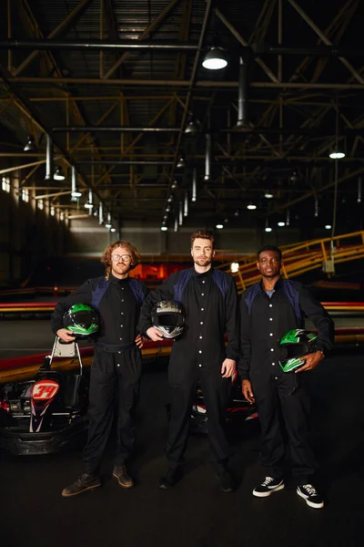Multicultural go kart drivers em ternos de proteção de pé e segurando capacetes, concorrentes — Fotografia de Stock