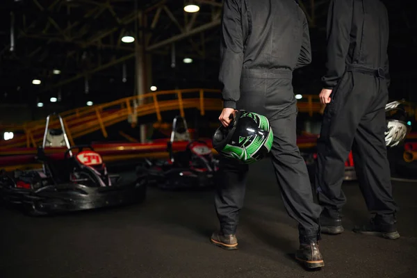 Cropped view of go kart drivers in sportswear walking on circuit and holding helmets, competitors — Stock Photo
